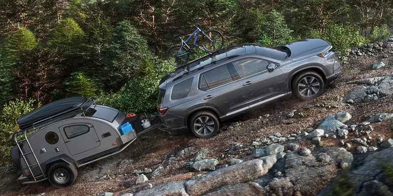 A gray 2024 Honda Pilot driving uphill on a rocky mountain road, towing a camper.