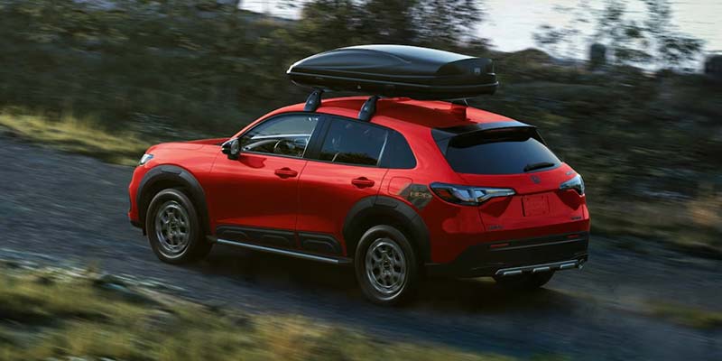 The image shows a red Honda HR-V SUV driving on a dirt road with a roof cargo box.