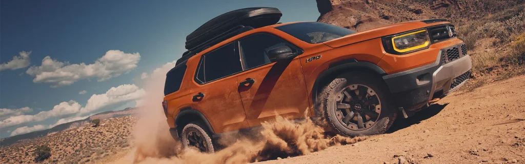 The image shows an orange Honda Passport SUV driving off-road, kicking up dust in a rugged terrain.
