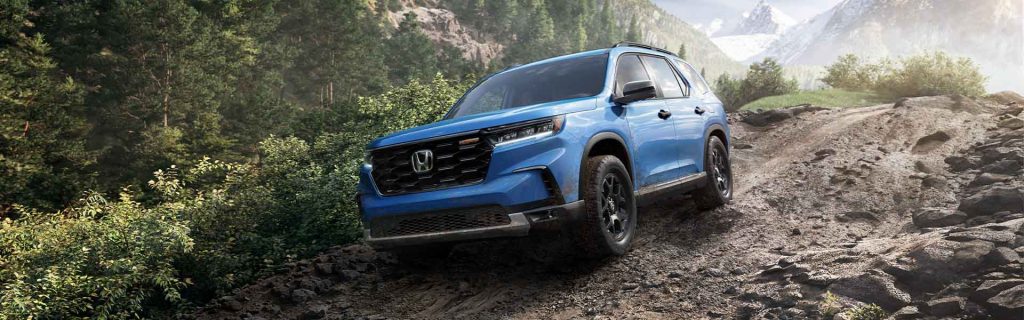 The image shows a blue Honda Pilot SUV driving on a rocky, off-road terrain surrounded by trees and mountains.
